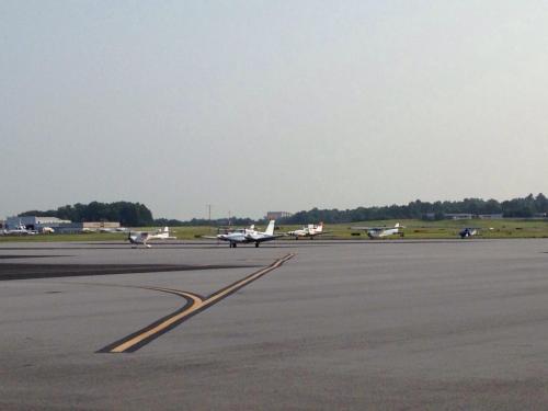 A view of a small airport runway with several light aircraft parked and taxiing on the tarmac.