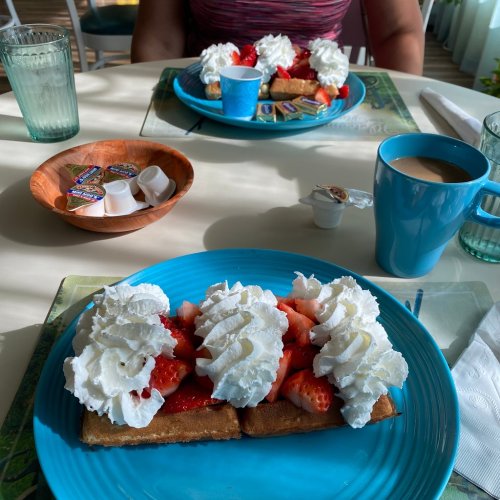 Two plates of waffles topped with strawberries and whipped cream, with coffee and condiments on a table.