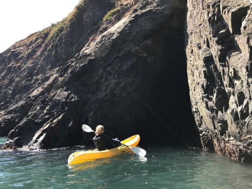 A person in a yellow kayak paddles near a rocky coastline and a dark cave entrance. Sunlight reflects on the water.
