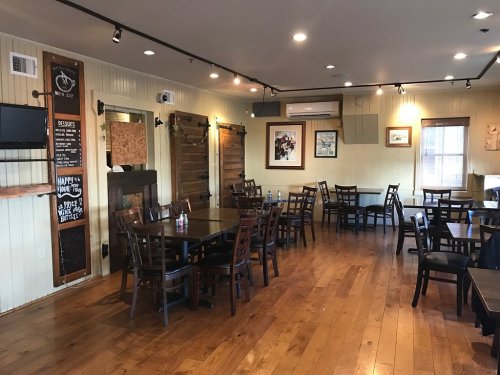 Interior of a restaurant with wooden floors, empty tables, and a chalkboard menu on the wall.