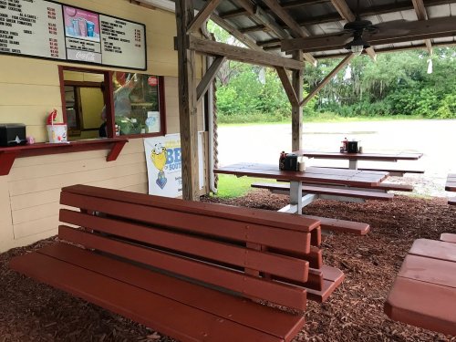 Outdoor seating area with wooden benches and tables, near a food counter under a covered structure. Green trees in the background.