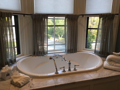 A luxurious bathtub by a large window, with elegant curtains and a view of greenery outside. Soft towels are nearby.