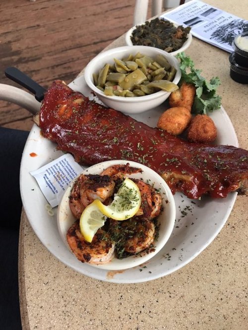 A plate featuring BBQ ribs with sauce, grilled shrimp with lemon, green beans, and hushpuppies.