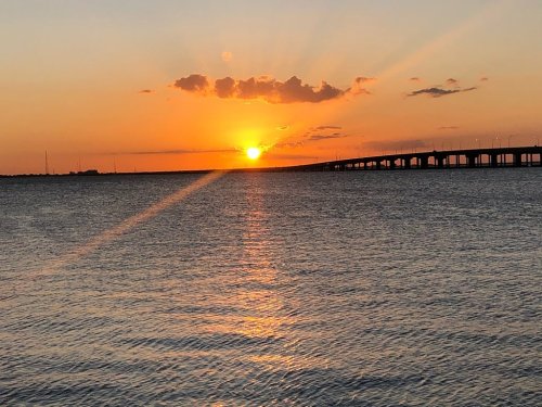 A vibrant sunset over a calm body of water, with rays of light and a bridge silhouetted in the background.