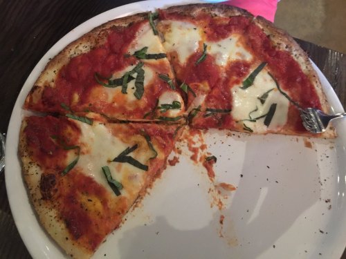 A partially eaten pizza with tomato sauce, mozzarella cheese, and fresh basil on a white plate.