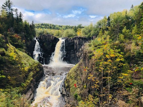 A scenic waterfall cascades between rocky cliffs, surrounded by lush greenery and autumn-colored trees.