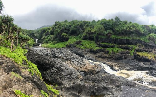 A lush, rocky landscape with waterfalls cascading down cliffs, surrounded by greenery under a cloudy sky.