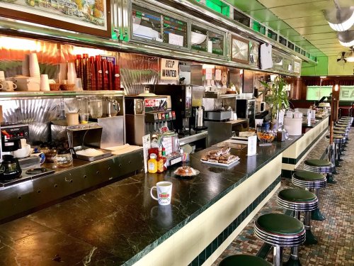 A retro diner interior featuring a long counter with stools, coffee mugs, and a variety of condiments and decor.