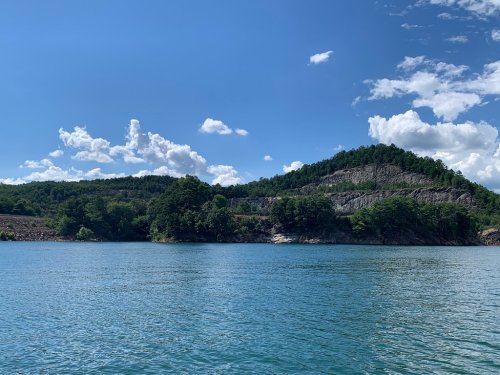 A serene lake surrounded by lush green hills and a clear blue sky with fluffy white clouds.