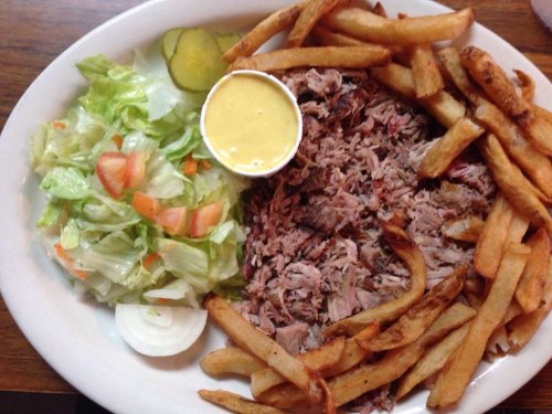 A plate of pulled pork, crispy fries, fresh salad with tomatoes, onions, and pickles, served with a side of yellow sauce.