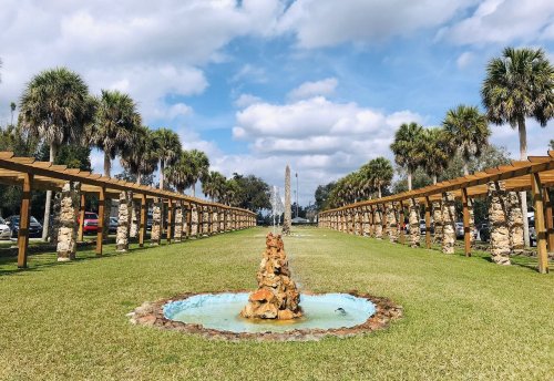A landscaped park featuring a central fountain, palm trees, and stone columns under a blue sky with clouds.