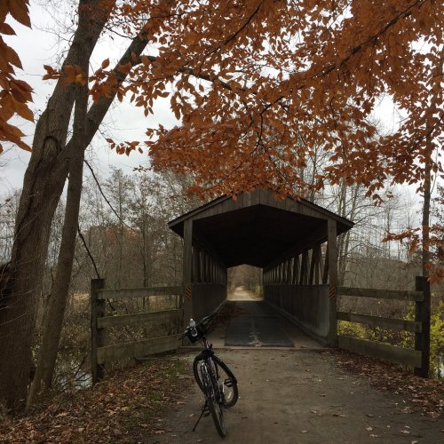 A bike rests beside a covered bridge surrounded by autumn leaves and trees on a quiet path.