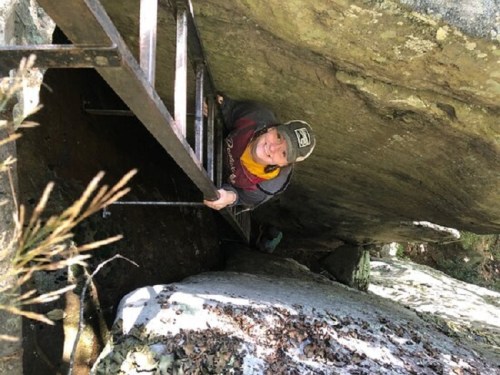 A person climbing a metal ladder against a rock formation, surrounded by nature.