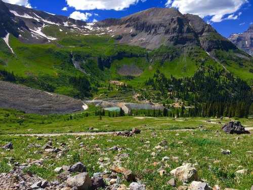 A scenic mountain landscape with green hills, rocky terrain, and a small pond surrounded by trees under a blue sky.