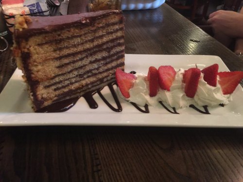 A slice of layered chocolate cake on a plate, accompanied by fresh strawberries and whipped cream drizzled with chocolate.