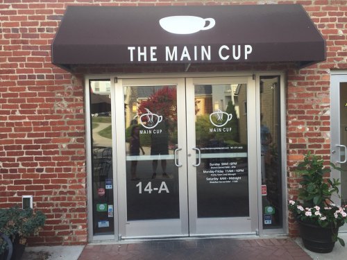 Entrance of "The Main Cup" café with glass doors and a brick wall, featuring a coffee cup logo above.