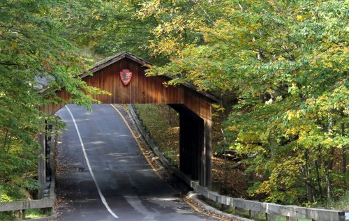 A wooden covered bridge with a park sign, surrounded by vibrant autumn foliage and a winding road.