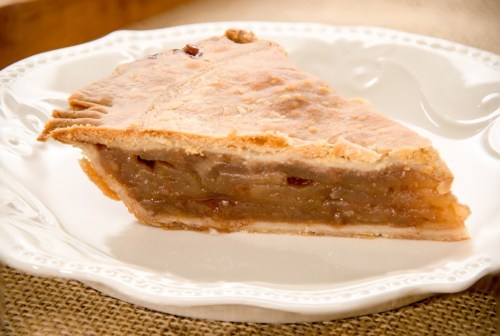 A slice of apple pie on a decorative white plate, showcasing a golden crust and a rich, apple-filled interior.