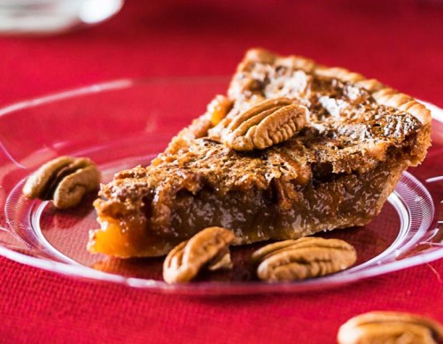 A slice of pecan pie on a clear plate, garnished with whole pecans, set against a red tablecloth.