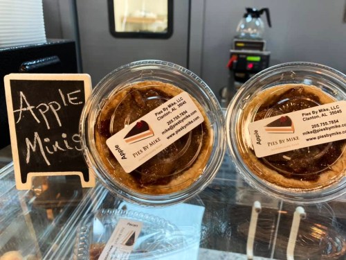 Two mini apple pies in clear containers, displayed behind a small chalkboard sign that reads "Apple Minis."