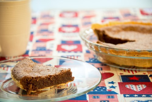 A slice of chocolate pie on a glass plate next to a whole pie, set on a patterned tablecloth.