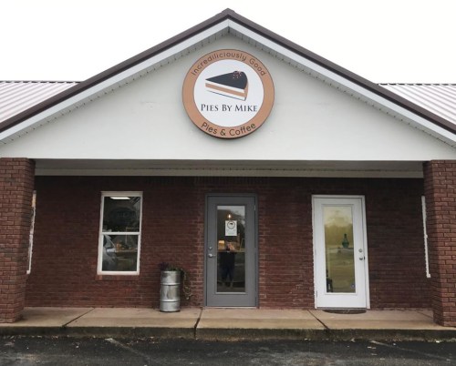 Exterior of "Pies by Mike" shop featuring a round sign and two entrance doors, with a brick facade and metal roof.