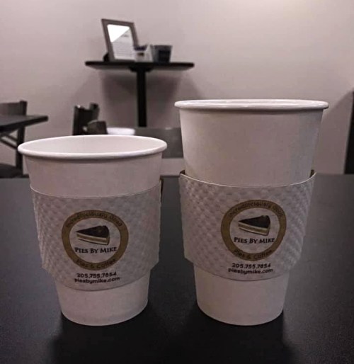 Two coffee cups, one small and one large, both with "Pies By Mike" labels, on a black table in a cafe setting.