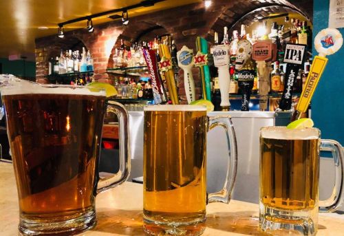 Three beer glasses of varying sizes sit on a bar, with a colorful selection of beer taps and bottles in the background.