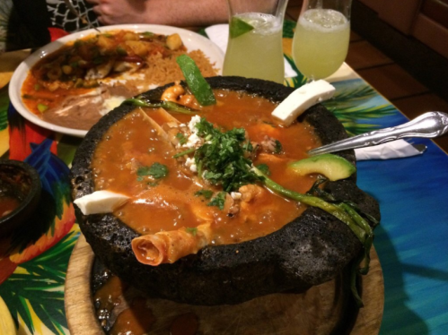 A vibrant dish served in a stone bowl, featuring a rich sauce, garnished with herbs and cheese, alongside drinks.