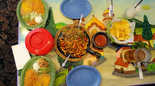 A colorful table spread with sizzling fajitas, rice, beans, guacamole, chips, salsa, and drinks.
