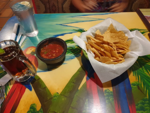 A colorful table with a bowl of salsa, a basket of tortilla chips, and a glass of iced tea.