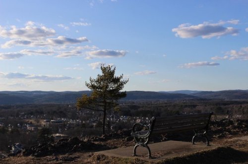 A scenic view from a hilltop bench overlooking a valley with mountains and a blue sky dotted with clouds.
