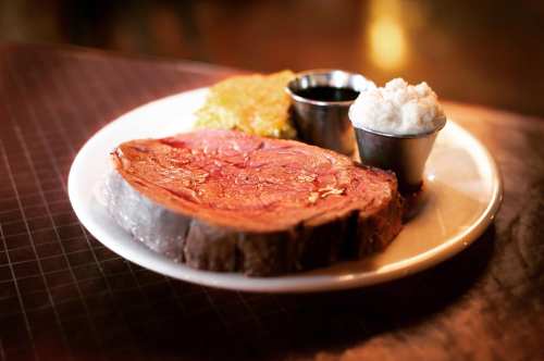 A plate featuring a large slice of prime rib, mashed potatoes, corn, and a small cup of sauce.