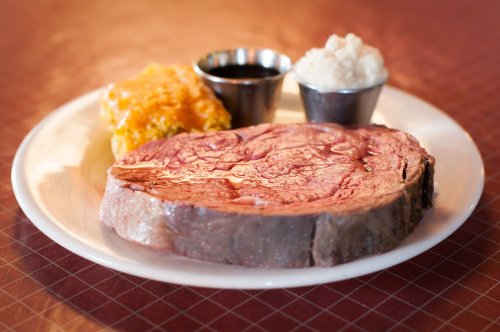 A plate featuring a slice of prime rib, cheesy casserole, and two small cups of sauce and mashed potatoes.