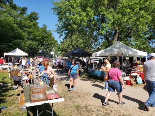 A bustling outdoor market with vendors, tents, and visitors browsing various goods under a clear blue sky.