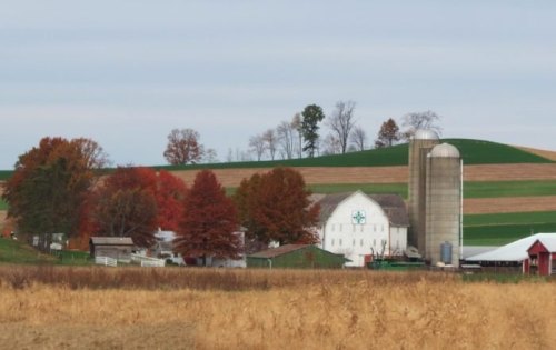 A scenic farm landscape with a white barn, silos, and colorful autumn trees against rolling green hills.
