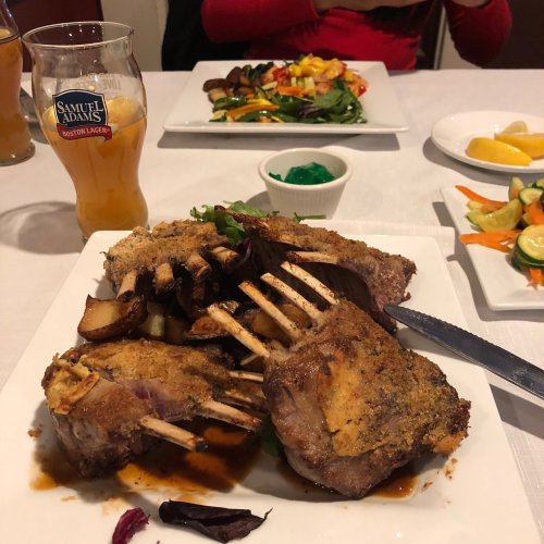 A plate of crusted lamb chops with potatoes, accompanied by a glass of beer and colorful side dishes in the background.