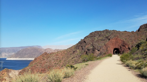 A scenic path leads to a tunnel, with rocky hills and a lake in the background under a clear blue sky.