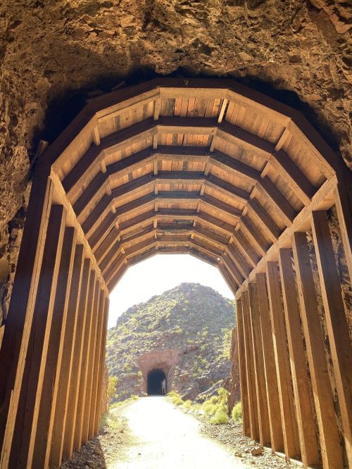 A wooden tunnel entrance leads to a rocky path, with another tunnel visible in the distance. Sunlight illuminates the scene.
