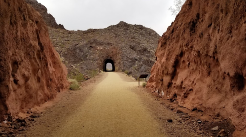 A dirt path leads through a rocky tunnel, flanked by reddish-brown cliffs under a cloudy sky.