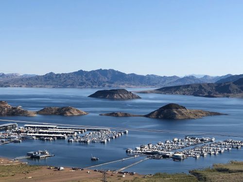 A scenic view of a marina with boats, surrounded by calm water and mountains under a clear blue sky.