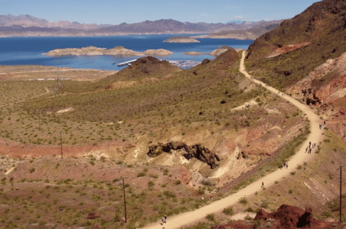 A scenic view of a winding dirt path through arid hills, overlooking a lake and distant mountains under a clear blue sky.