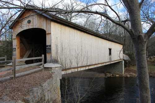 A wooden covered bridge spans a river, surrounded by bare trees and a cloudy sky.