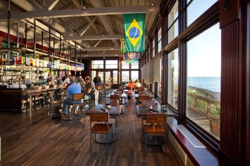 Interior of a restaurant with a bar, wooden floors, and a Brazilian flag, overlooking a scenic waterfront view.