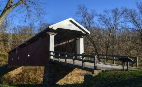 A red covered bridge with a wooden walkway, surrounded by bare trees and a clear blue sky.