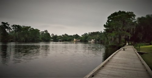 A serene lake scene with a wooden walkway, surrounded by trees and a cloudy sky, reflecting calm waters.