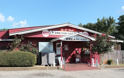 Exterior of Dreamland BBQ restaurant, featuring a red building, signage, and a clear blue sky.