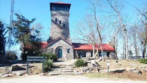 Historic stone building with a red roof and tower, surrounded by trees and rocky terrain under a clear blue sky.