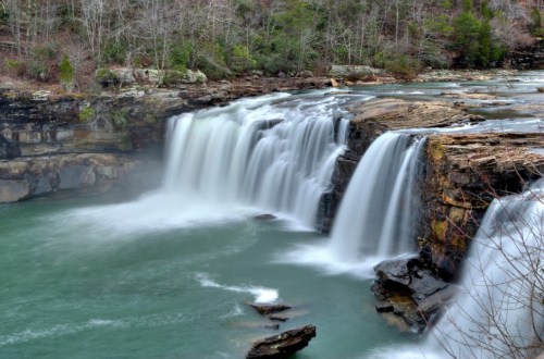 A serene waterfall cascades over rocky cliffs into a tranquil turquoise pool, surrounded by lush greenery.