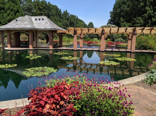 A serene garden scene featuring a pond with lily pads, colorful flowers, and a gazebo under a clear blue sky.